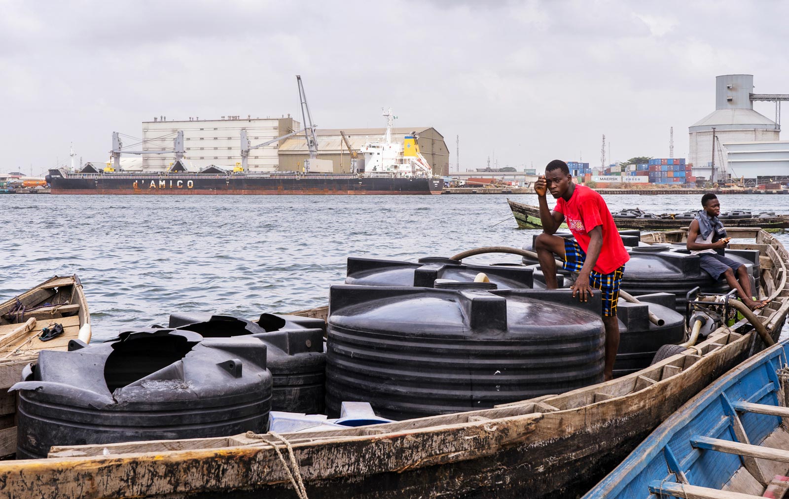 Two young men on a wooden boat at a dock pumping water out of large black water containers.