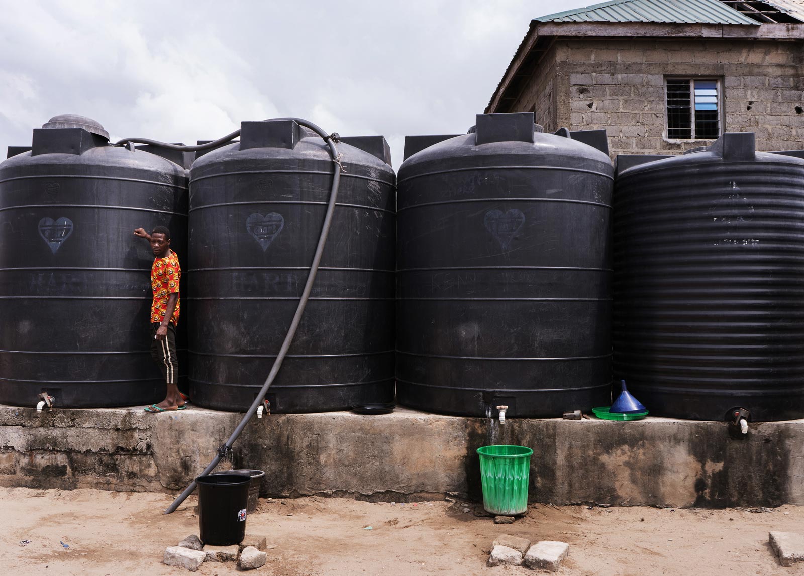 A young man standing in front of 4 large black water tanks that are nearly twice his height.