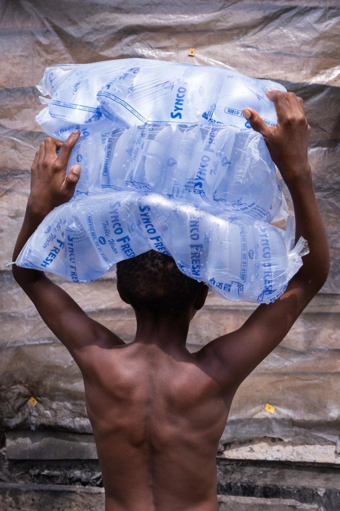 The back of a young boy balancing a pile of water sachets on his head.