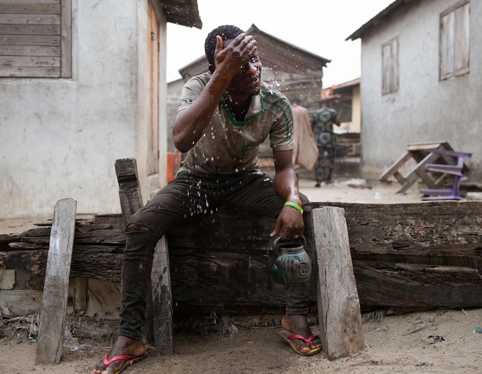 A man sitting down, washes his face using water from a small pot