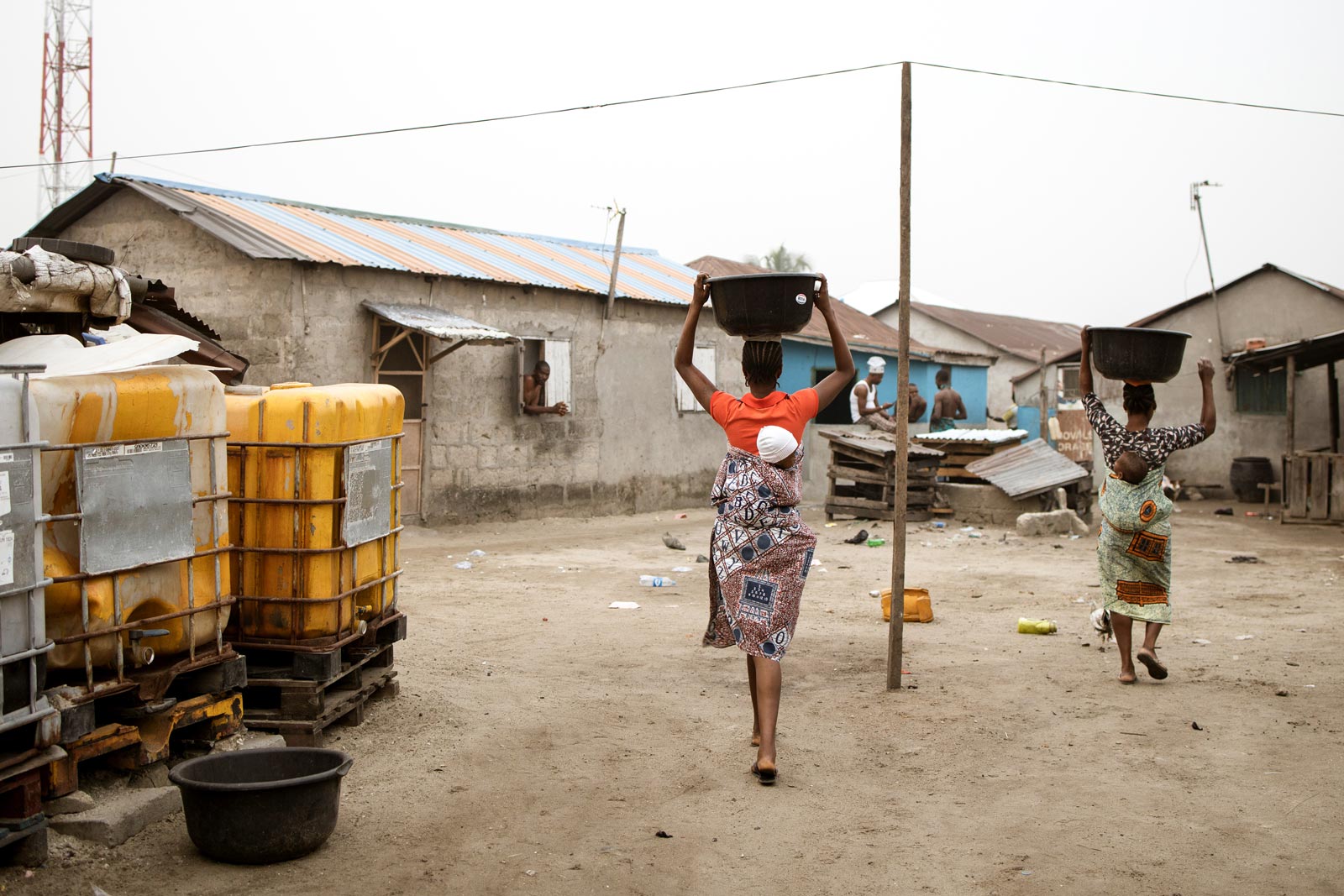 Two women, each carrying a baby on their back, balance buckets filled with water on top of their head as they walk past two large yellow water tanks.