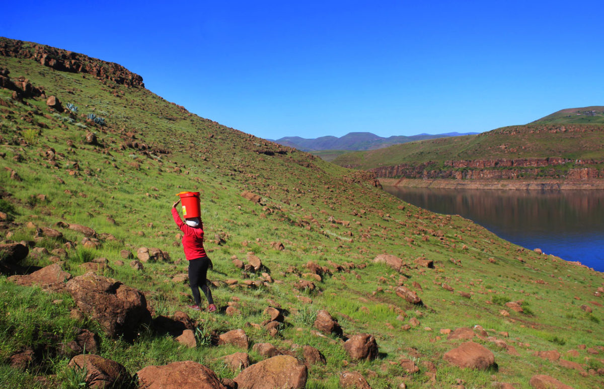 A young woman walks up a steep, rocky slope balancing a bucket of water on top of her head.